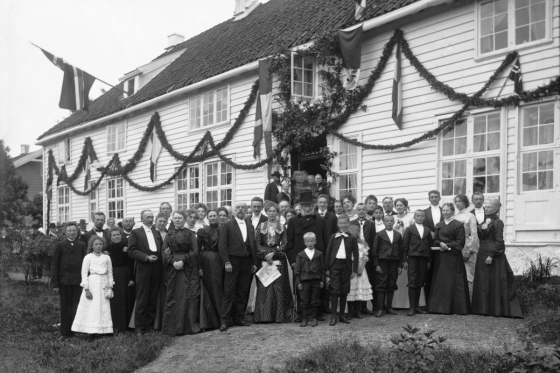 Fotografi i svart-hvitt som viser en stor gruppe festkledte mennesker foran en pyntet trebygning. Fotoet er fra 1904 og viser 100 års jubileet på Svanøy.