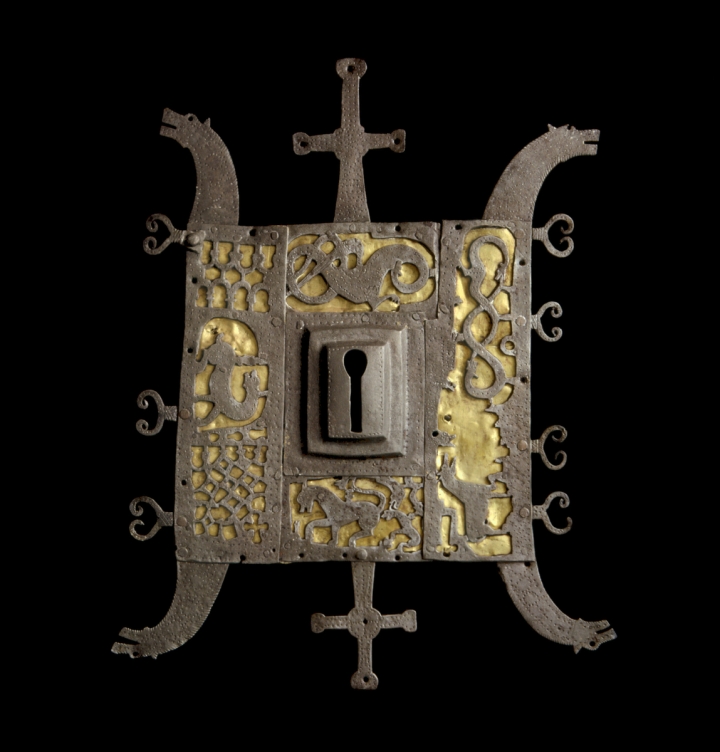 Gjenstandsfoto av et firkantet dørbeslag i jern med nøkkelhull. Dørbeslaget er dekorert med fire drakehoder i hver ende og gjennombrutte ornamenter i form av hester som ligger mot en gullfarget bunn.  